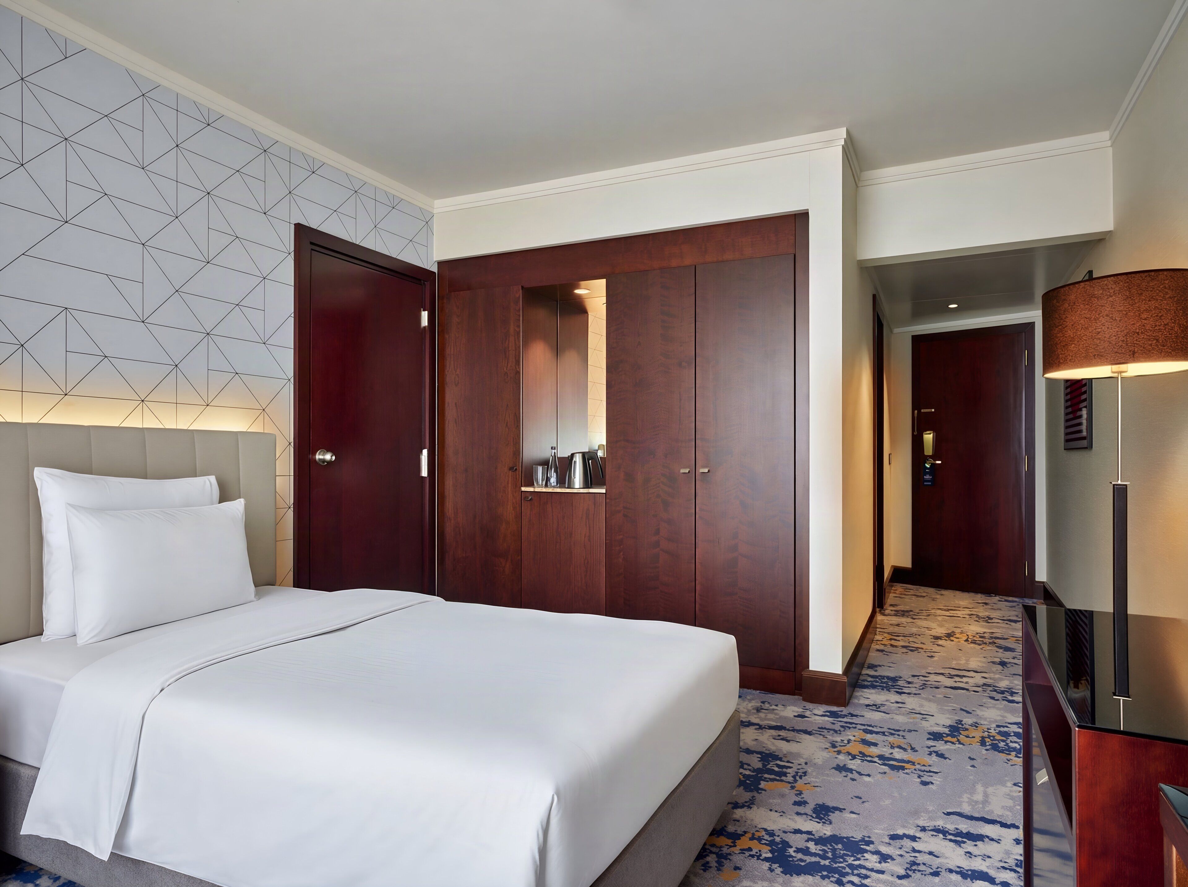 A hotel room with a bed, a wardrobe, a desk, and a lamp.