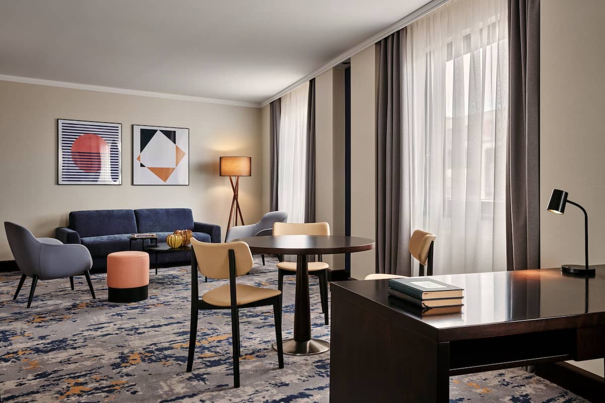 A hotel room with a sofa, chairs, a desk, a lamp, and artwork on the wall.