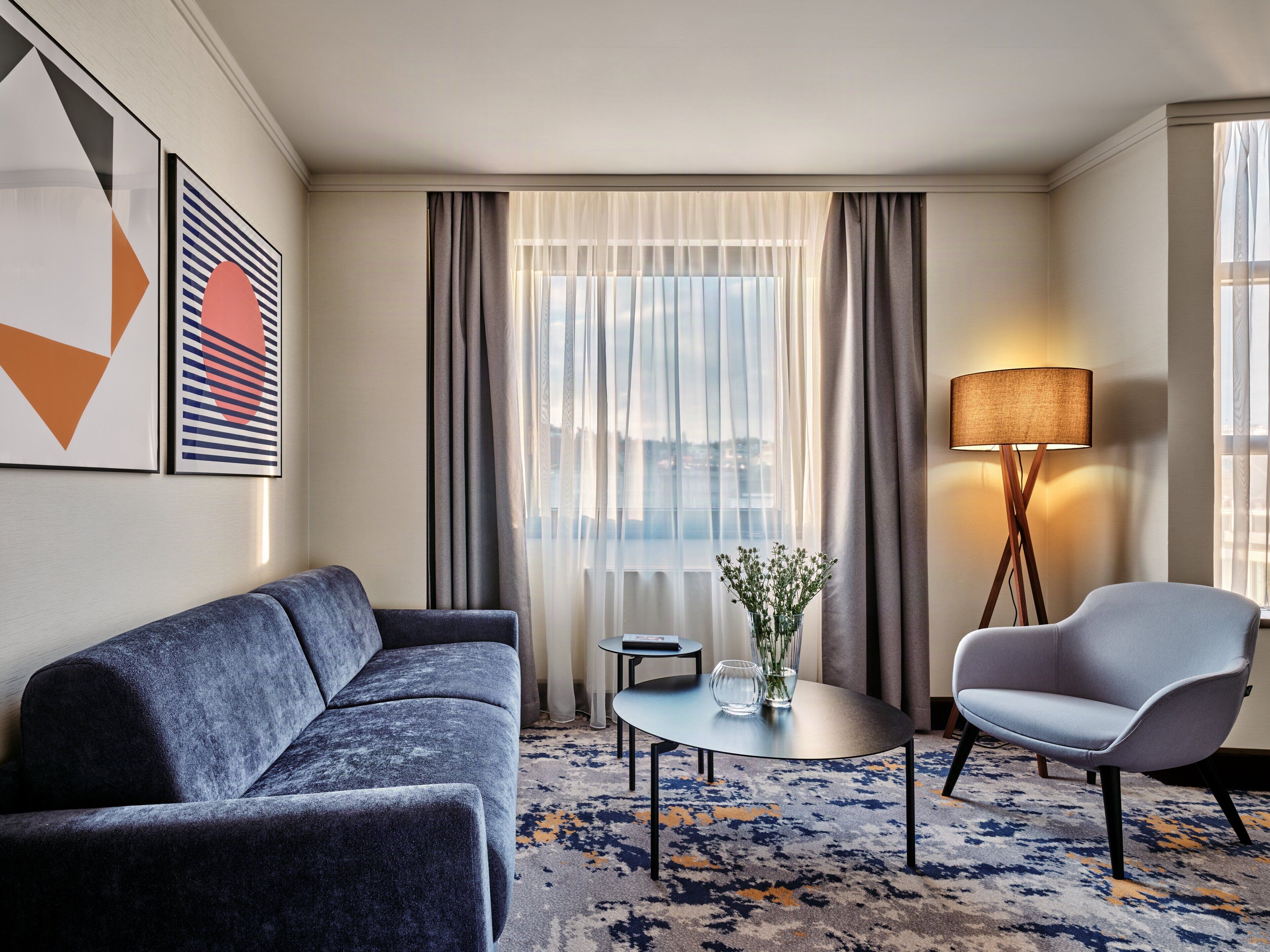 Een hotelkamer met een bank, fauteuil, klein tafeltje, lamp en uitzicht op de stad.