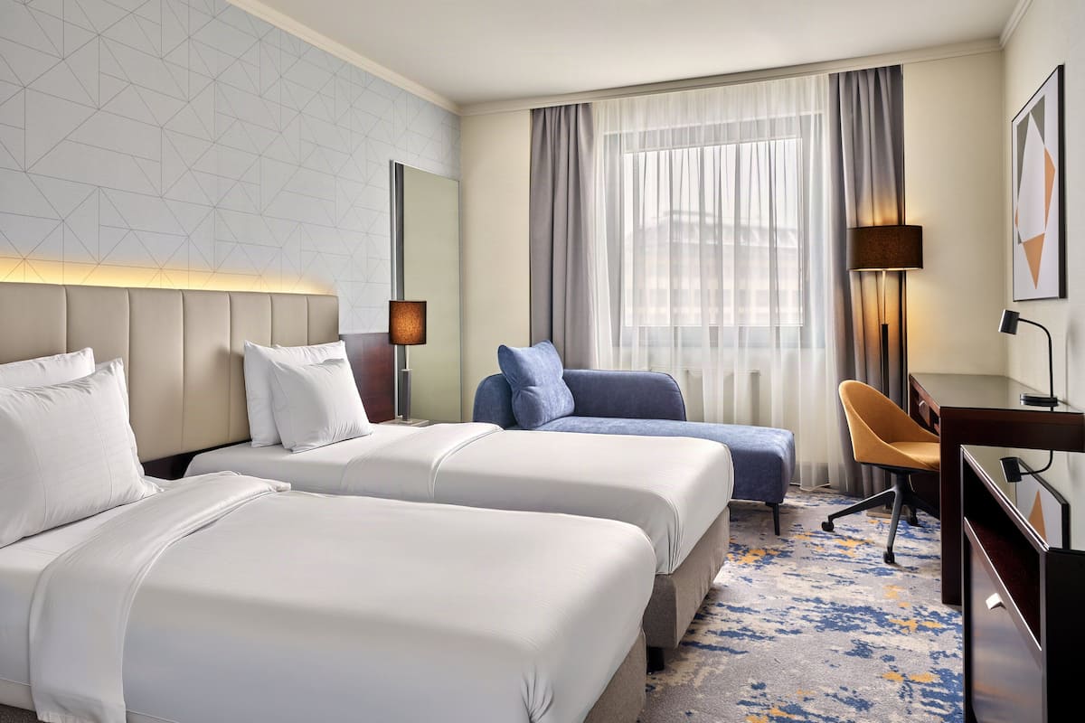 A hotel room with two beds, a desk, a chair, and a large window with curtains.