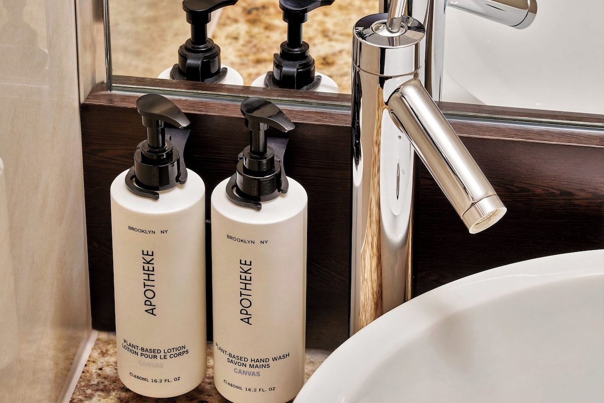 A bathroom with a sink, two bottles of Apothéke plant-based lotion, and a mirror reflecting a faucet.