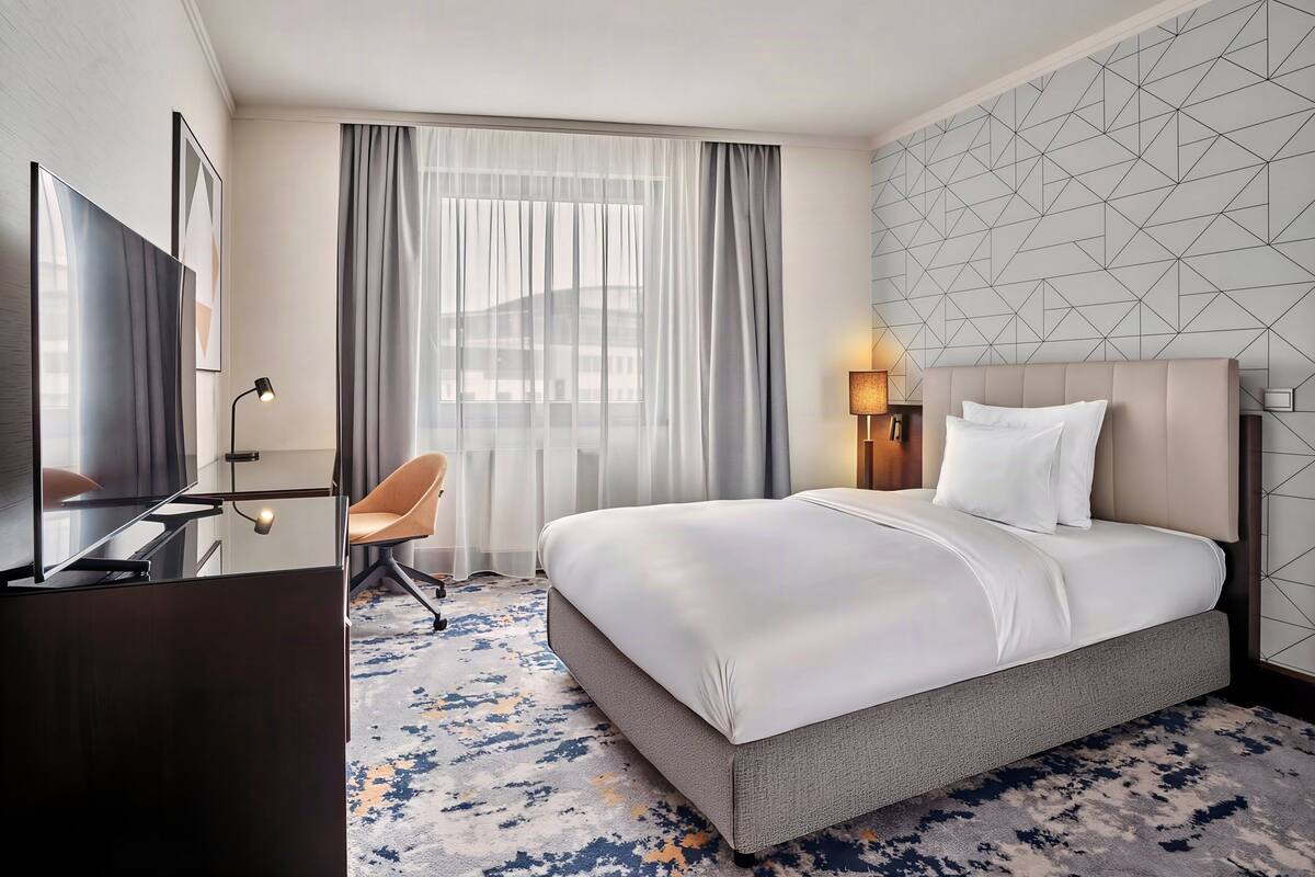 A hotel room with a bed, a desk with a TV, a chair, a window with curtains, and a geometric wall design.