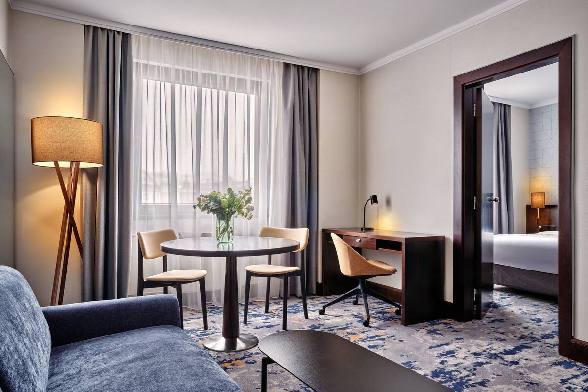 A hotel room with a round table, two chairs, a desk with a lamp, a bed, a large mirror, and a floor lamp.