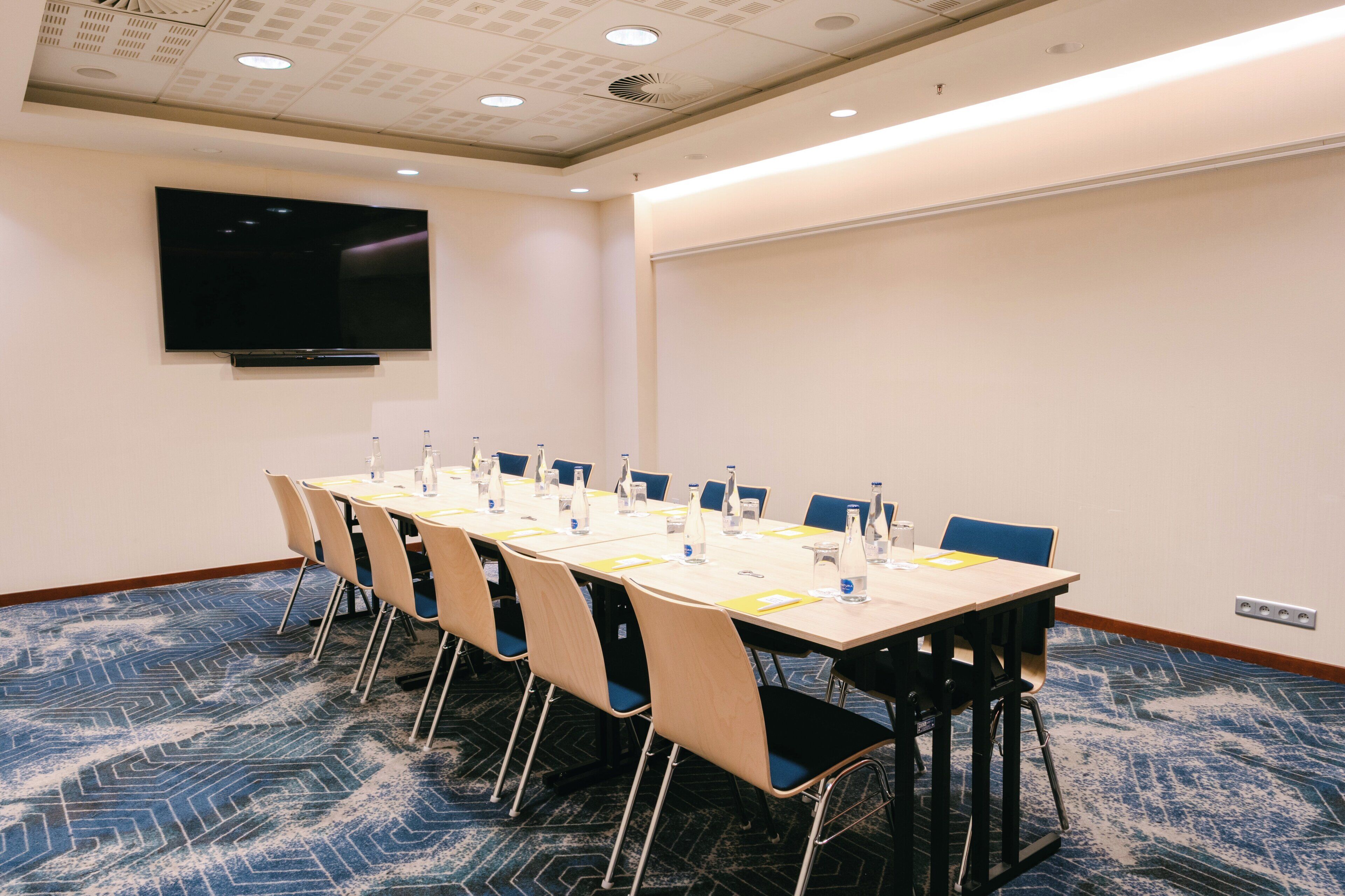 Un salón de conferencias con una larga mesa, sillas, botellas de agua y un televisor de pantalla plana en la pared.