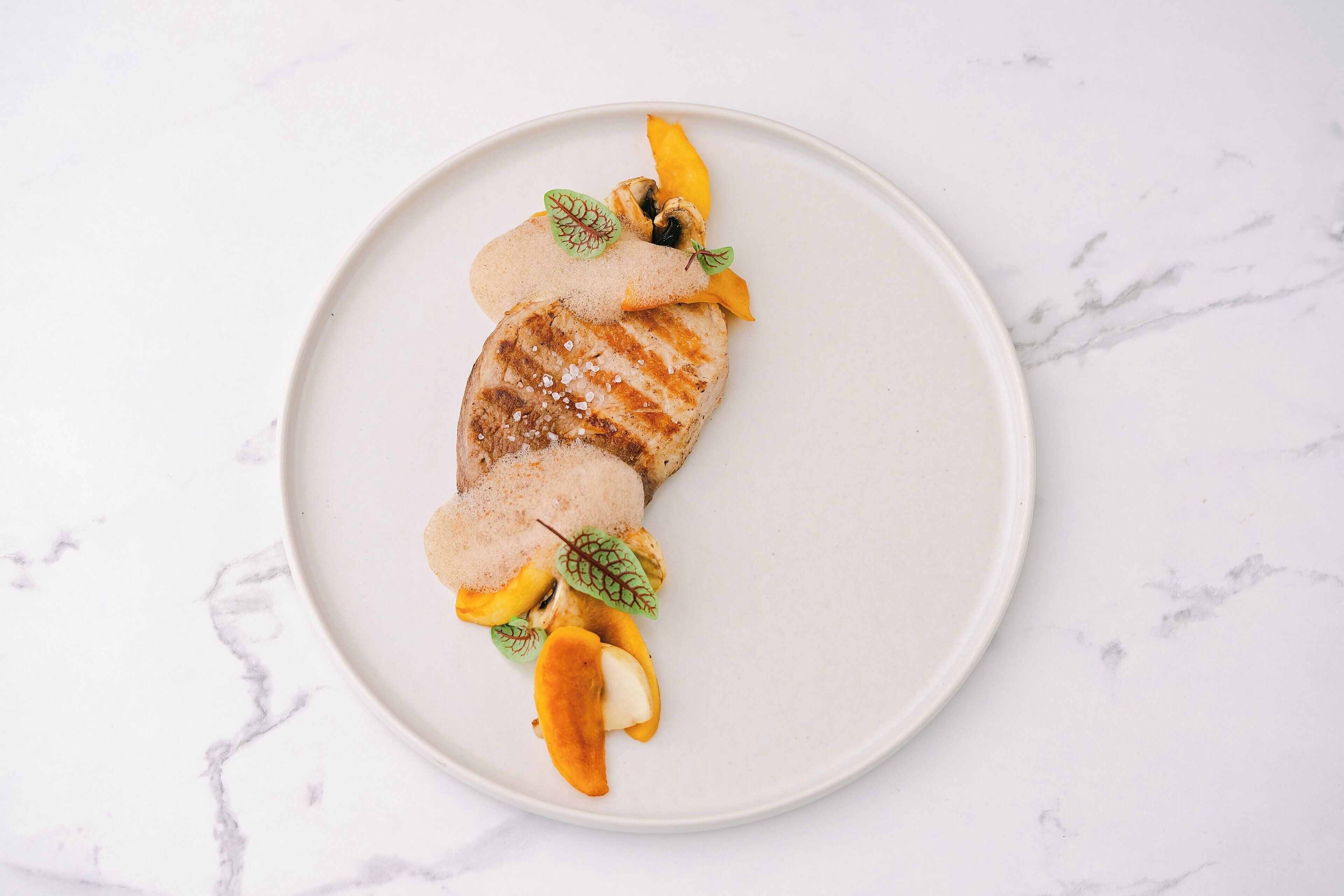 A plate with a grilled item, possibly chicken, accompanied by sliced mango and a green leaf garnish.