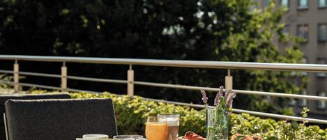 A table set with breakfast items including croissants, a bowl of fruit, and a glass of orange juice, with a cityscape and greenery in the background.