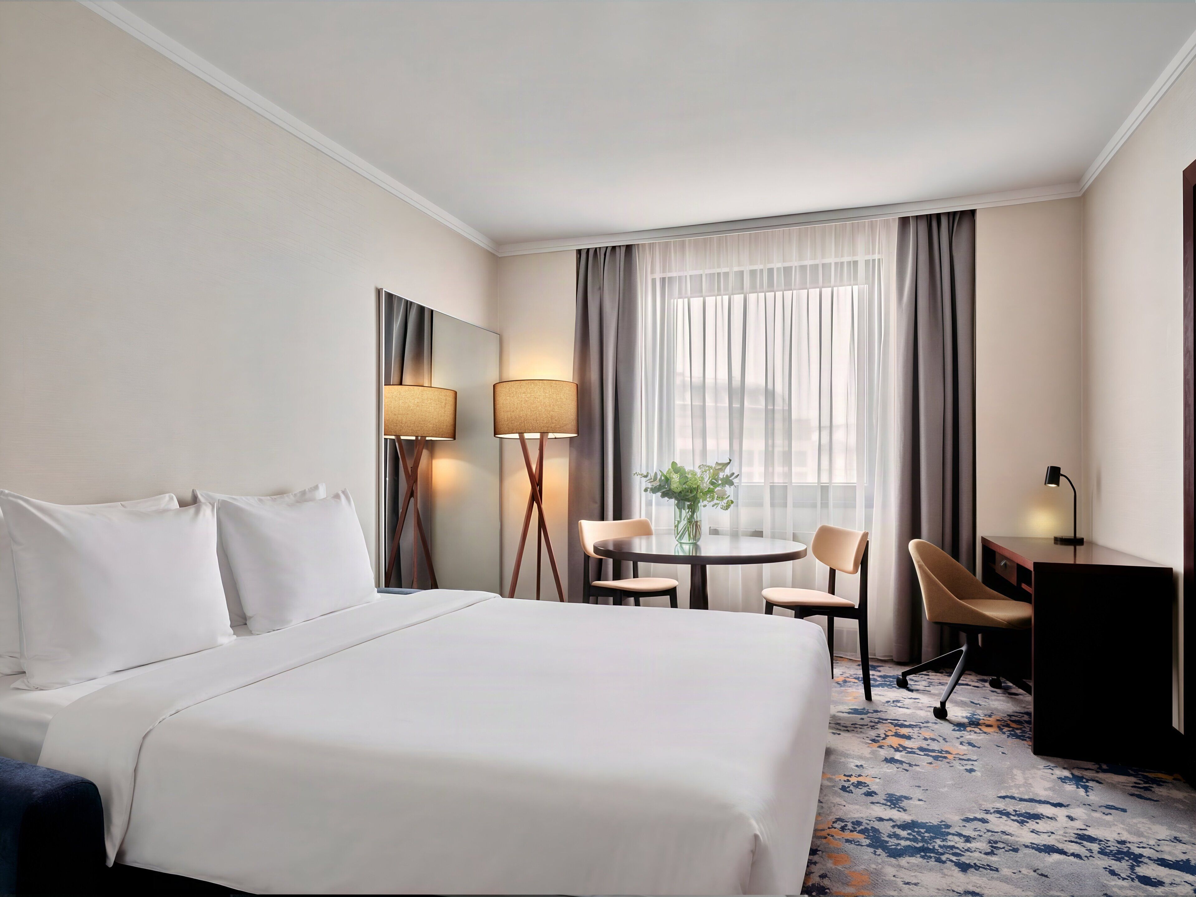 Een hotelkamer met een groot bed, een bureau, een stoel, een tafeltje met een vaas en een raam met gordijnen.