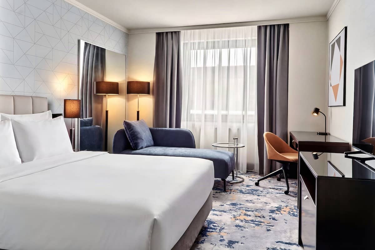 A hotel room with a bed, two chairs, a desk, a lamp, and a large window with curtains.