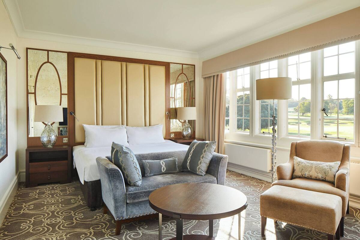 A hotel room with a large bed, two armchairs, a small round table, and a view of the outdoors through large windows.