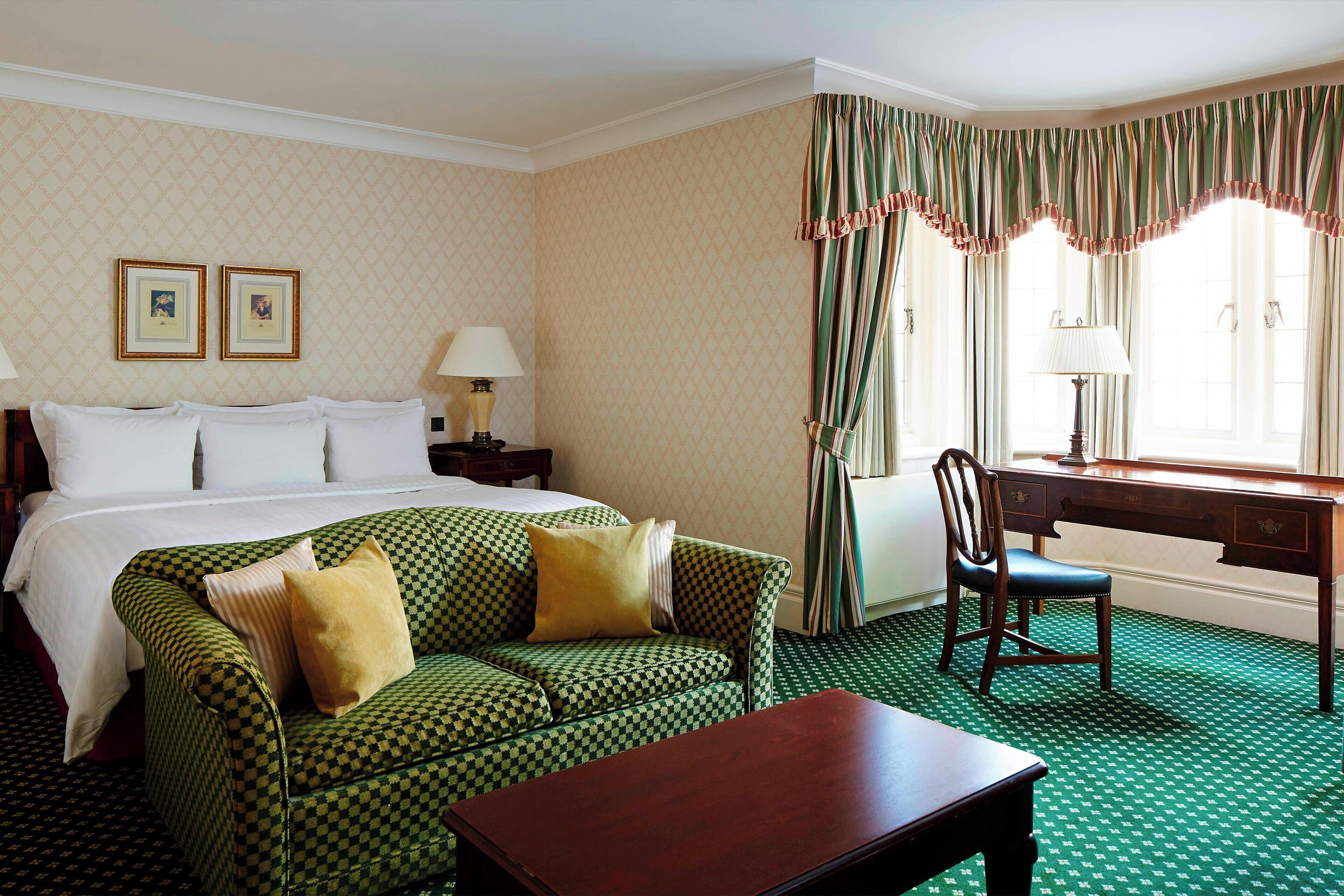 Una habitación de hotel con un sofá a cuadros, un escritorio de madera, una cama con sábanas blancas y una ventana con cortinas.