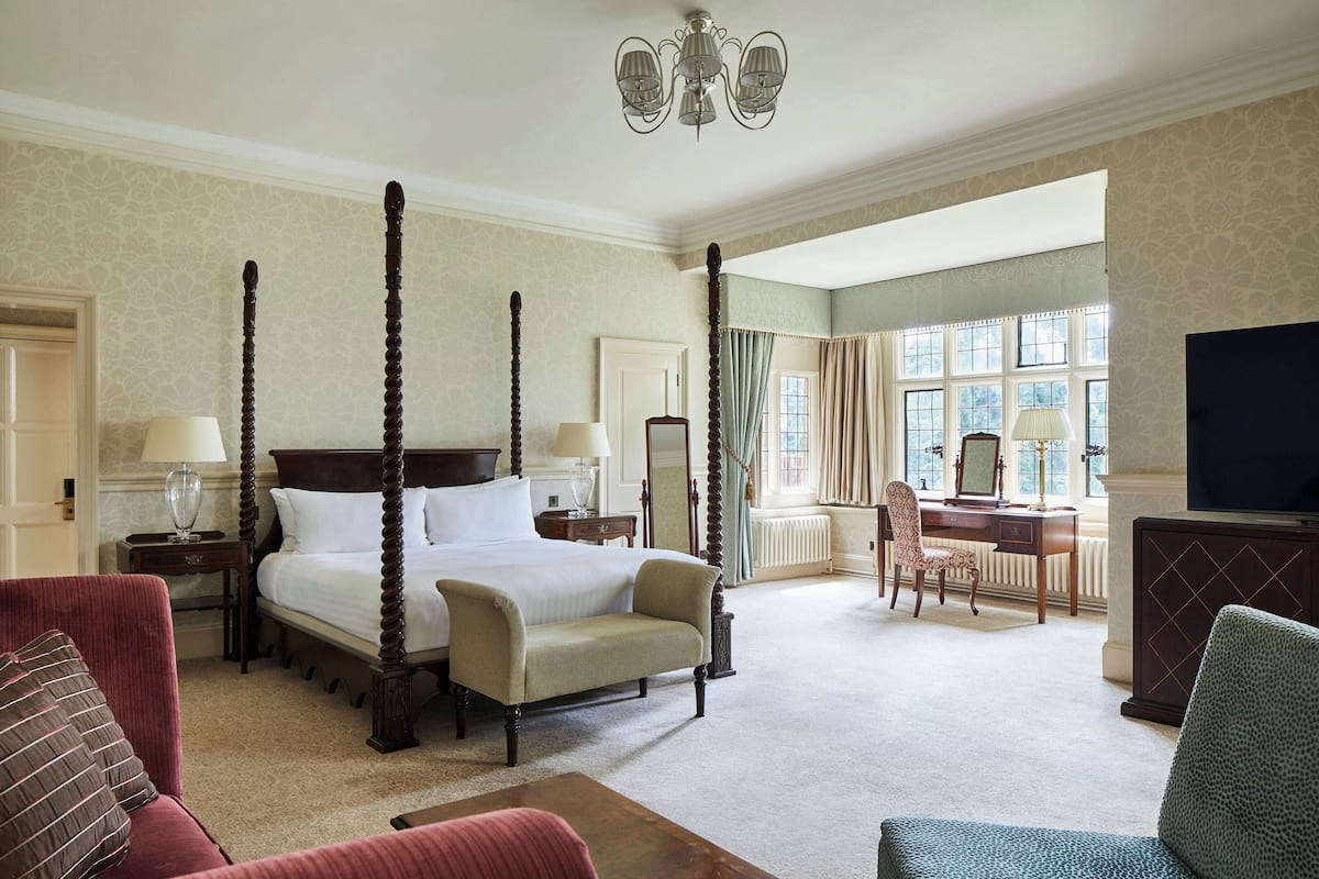 A four-poster bed with a wooden headboard, a sitting chair, a desk with a chair, and a television on a cabinet.