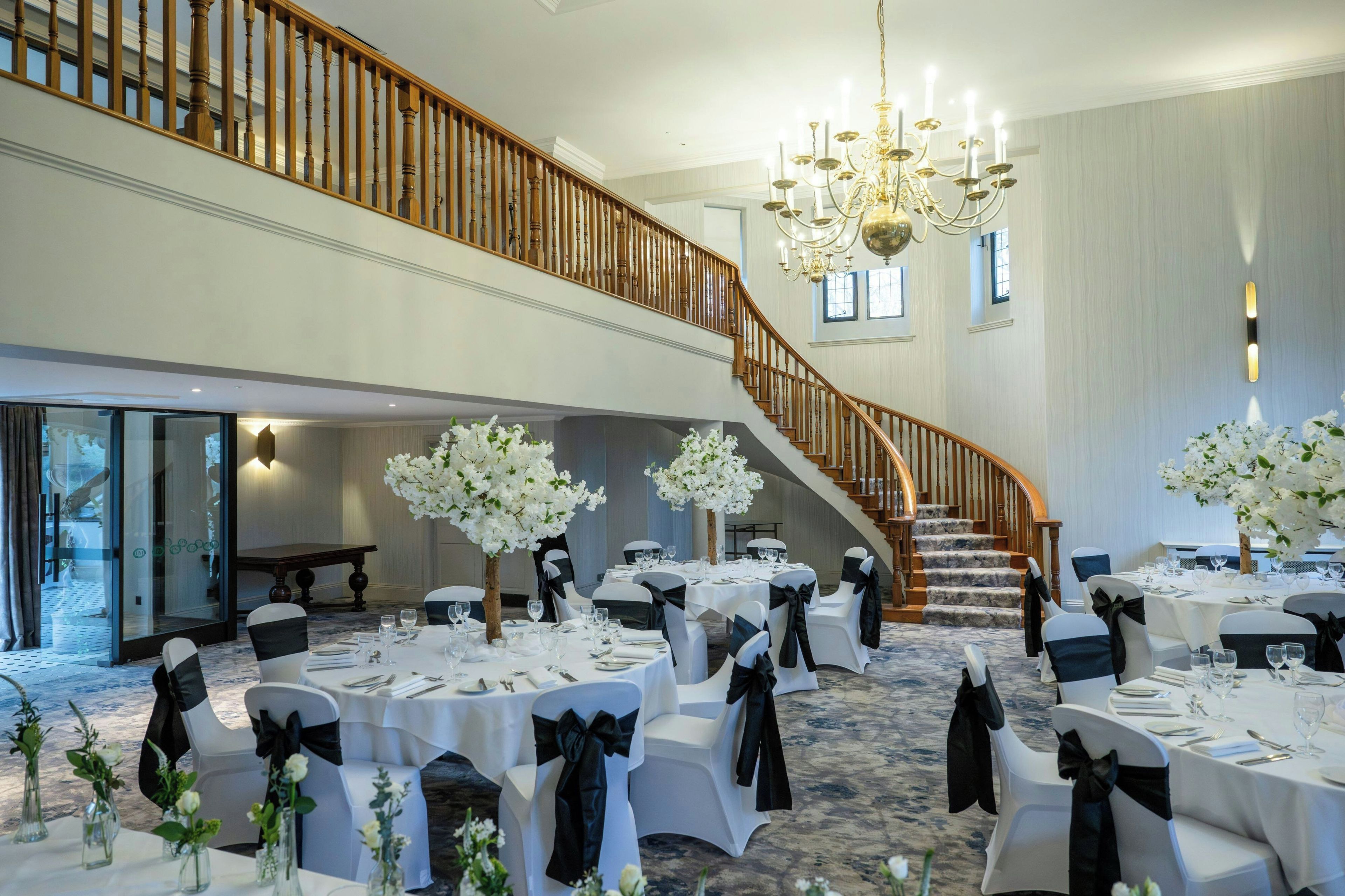 A spacious banquet hall with multiple round tables, each set with white tablecloths and black chair sashes. A grand staircase with wooden railings leads to an upper level.
