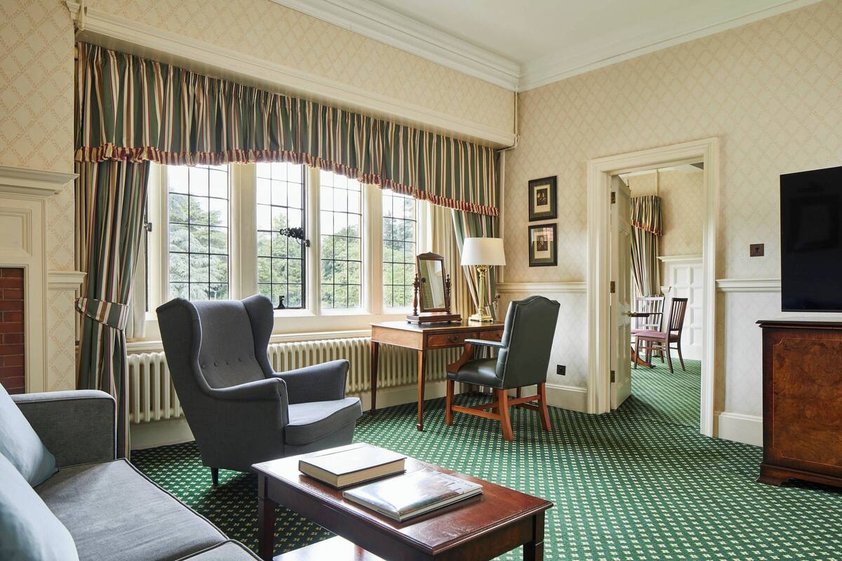 A room with a green patterned carpet, a fireplace, a sofa, a desk with a chair, and a television.
