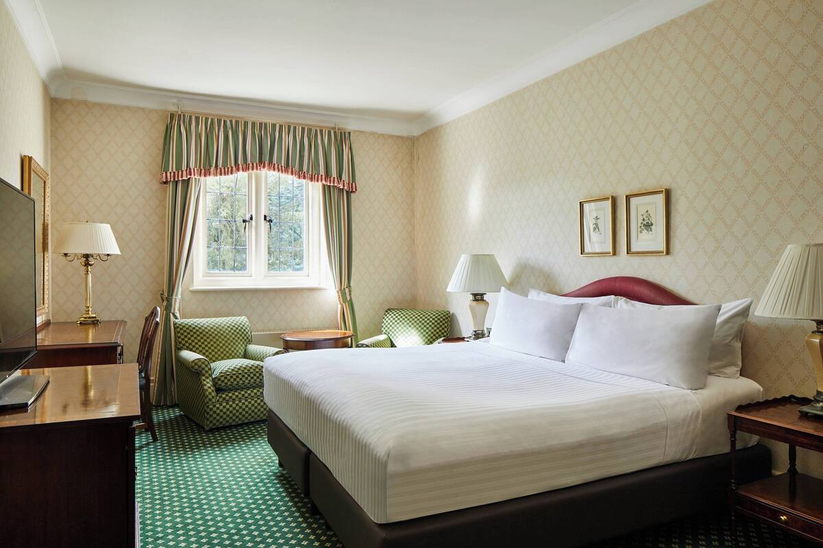 A hotel room with a large bed, a desk with a lamp, a chair, and a window with curtains.