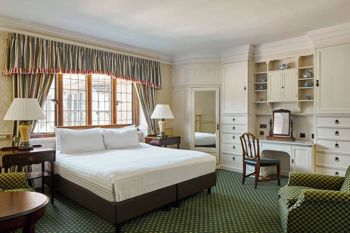 A hotel room with a large bed, two bedside tables with lamps, a desk with a chair, a wardrobe, and a mirror.