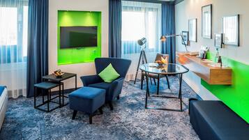 A modern hotel room with a flat-screen TV, a blue armchair, a glass coffee table, and a desk with a lamp.