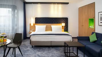 A hotel room with a bed, bedside tables, a desk, a chair, a sofa, and a window with curtains.