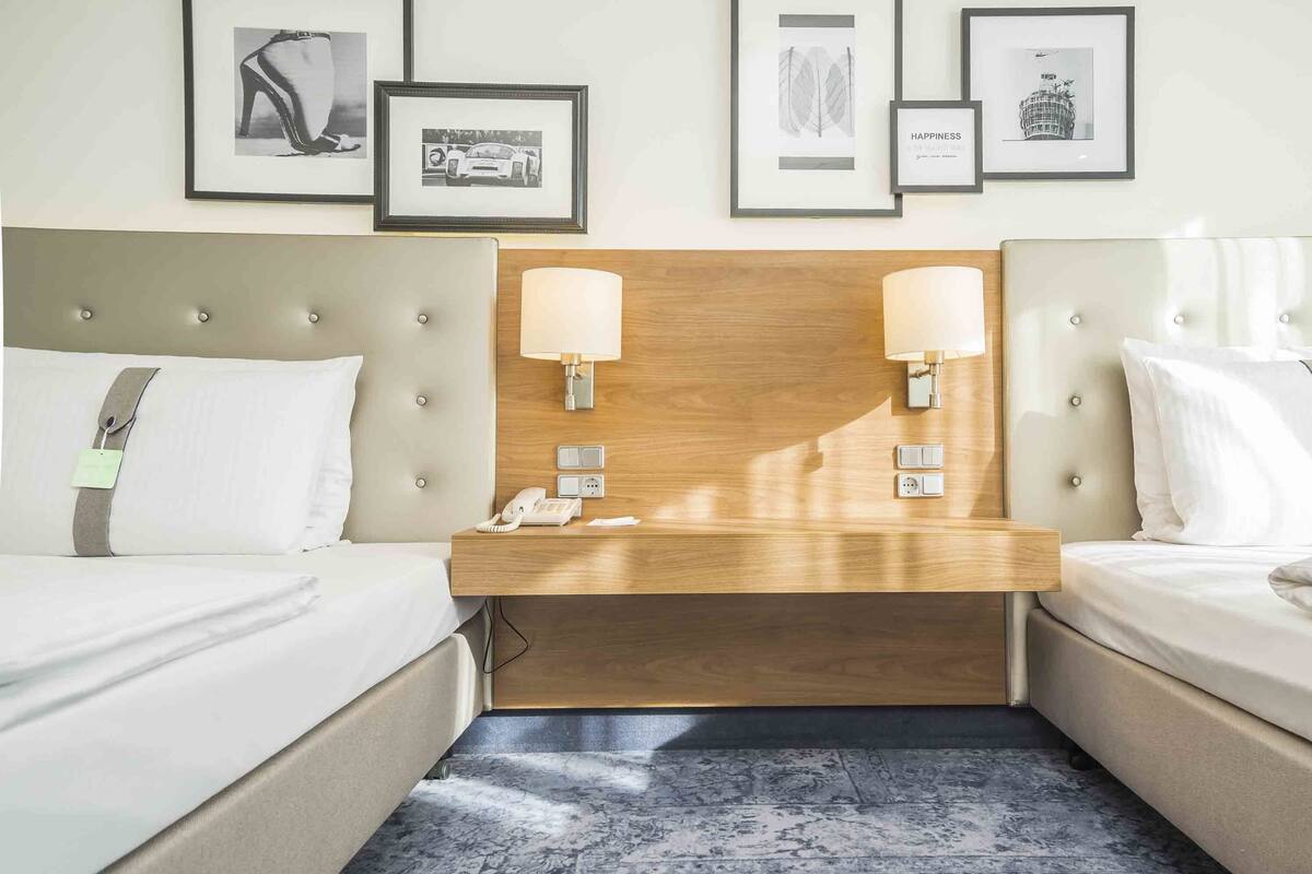 A hotel room with two beds, a wooden headboard, a nightstand with a phone and lamp, and framed pictures on the wall.