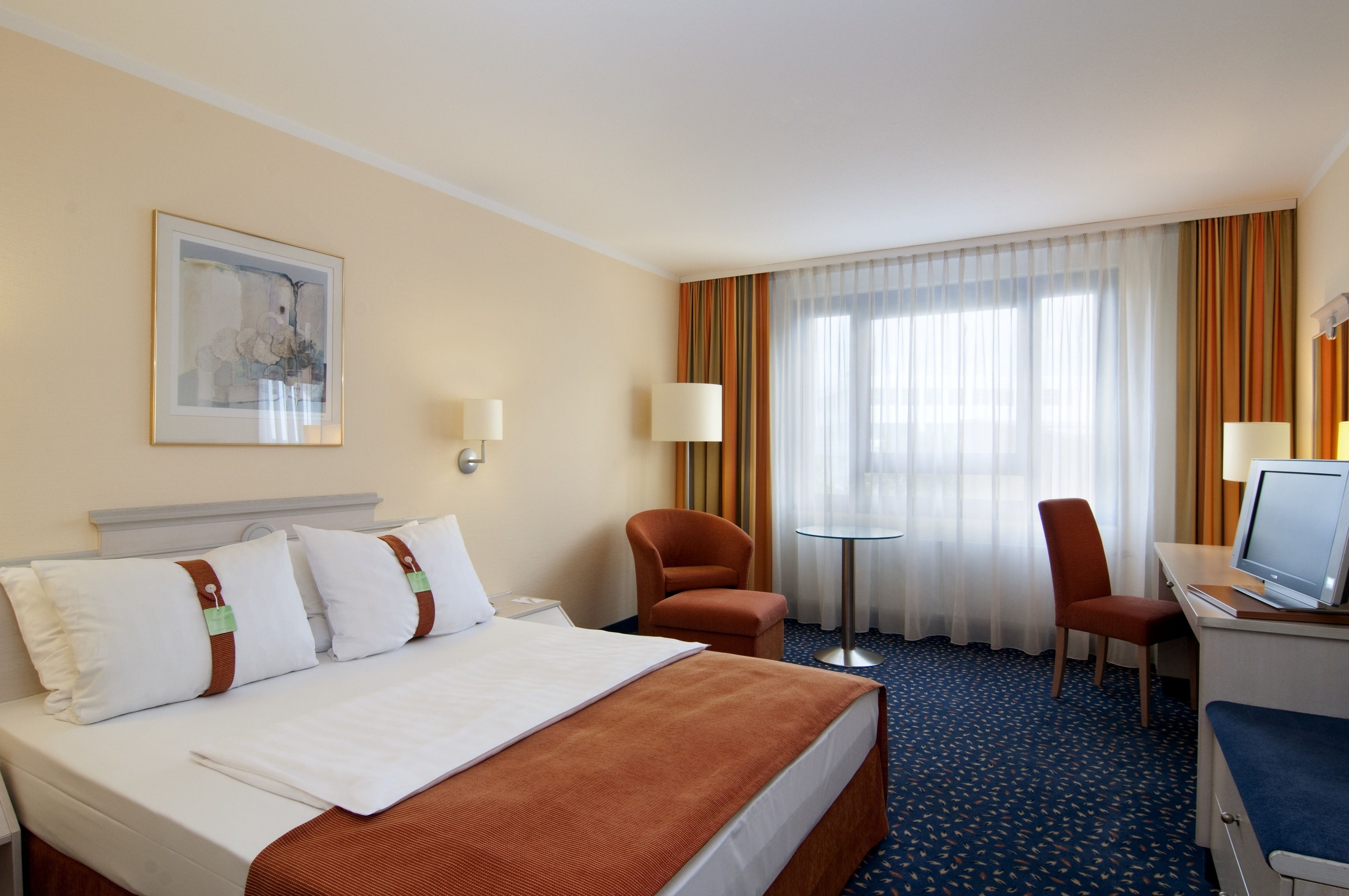 A hotel room with a bed, two chairs, a desk with a computer, and a window with curtains.