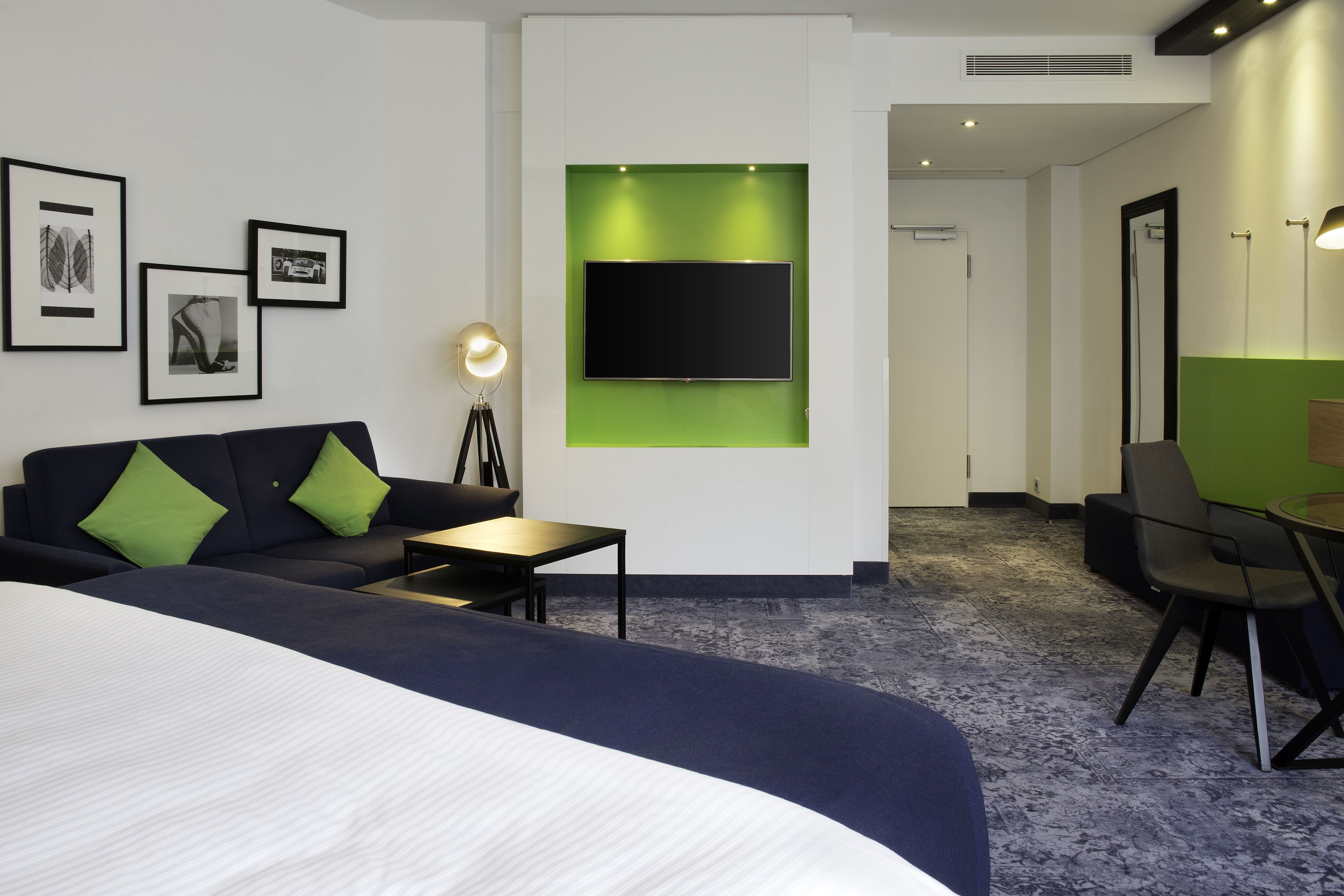 A modern hotel room with a large bed, a sofa, a desk, and a flat-screen TV mounted on a green accent wall.
