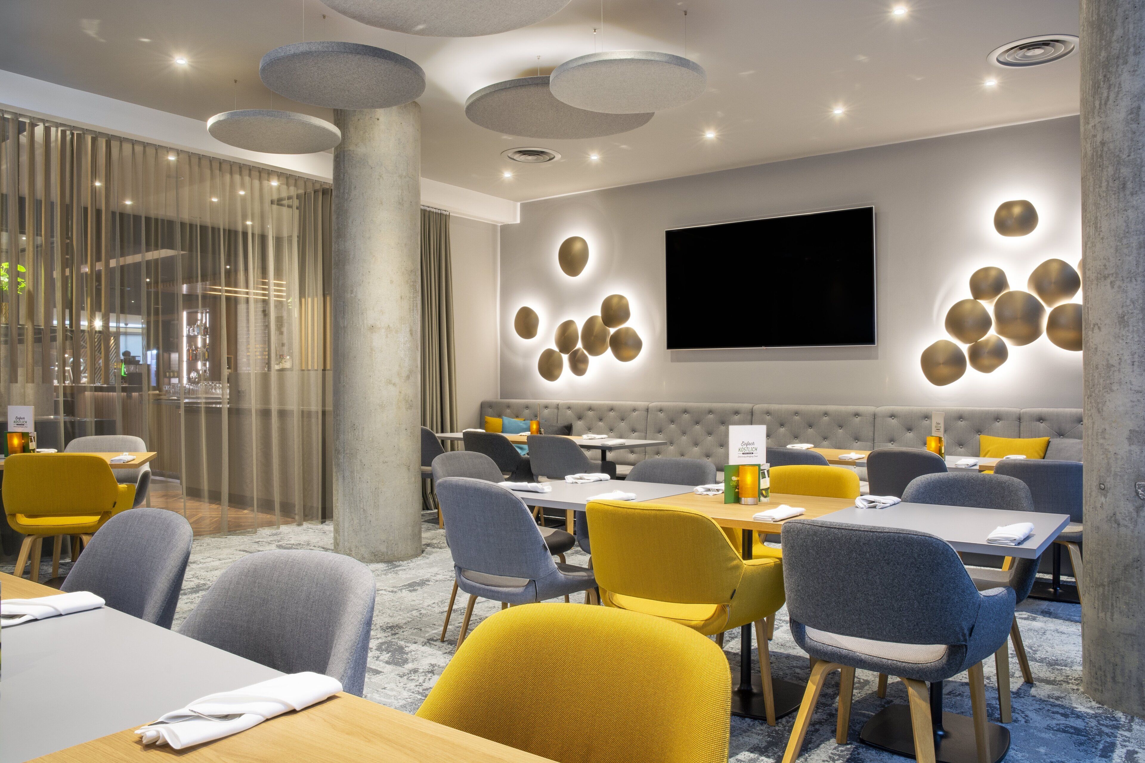 A modern dining area with round tables and chairs, a large flat-screen TV, and a seating area with a cushioned bench.