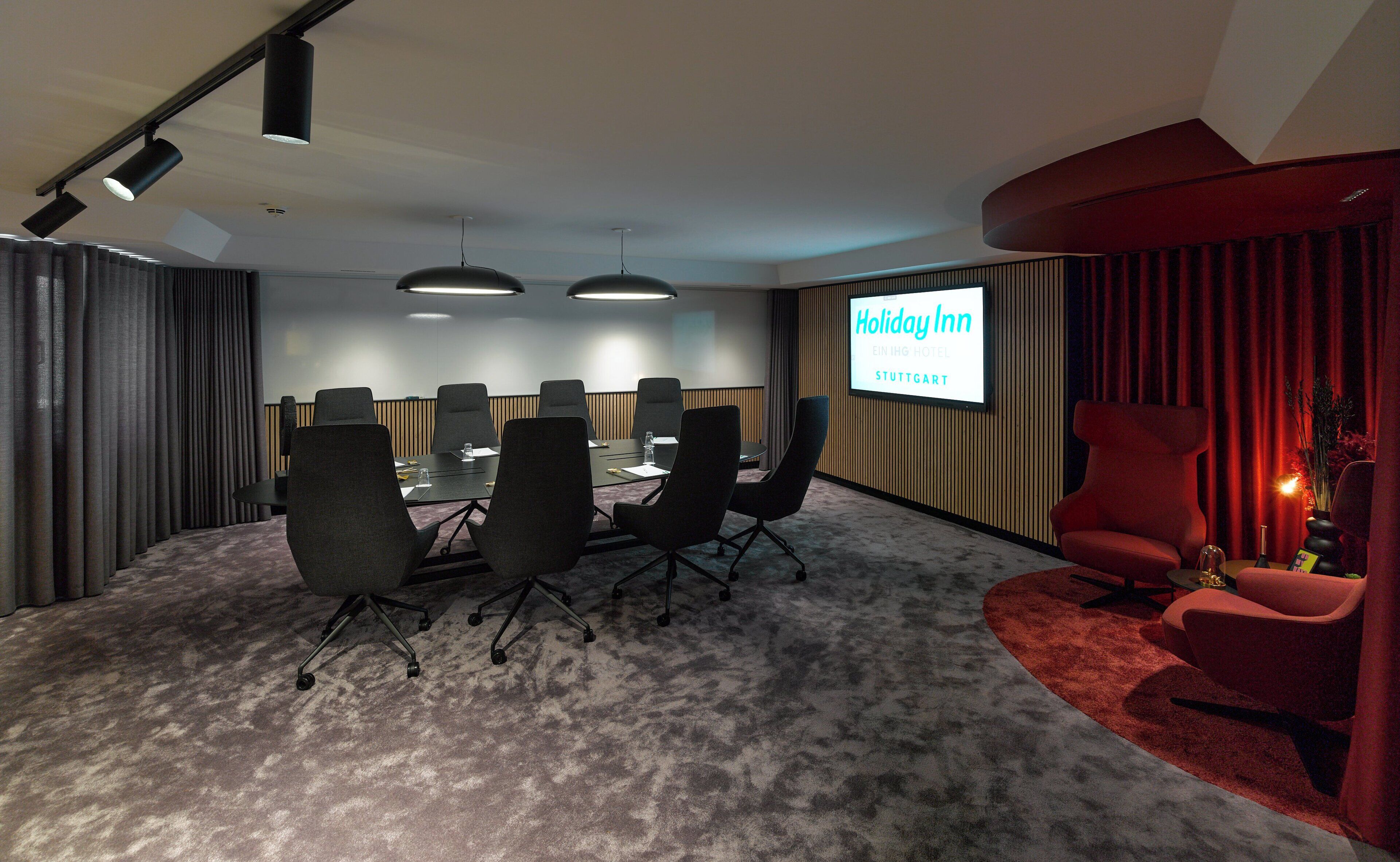 Un salón de conferencias con una gran mesa, sillas y una pantalla mostrando "Holiday Inn Stuttg..."