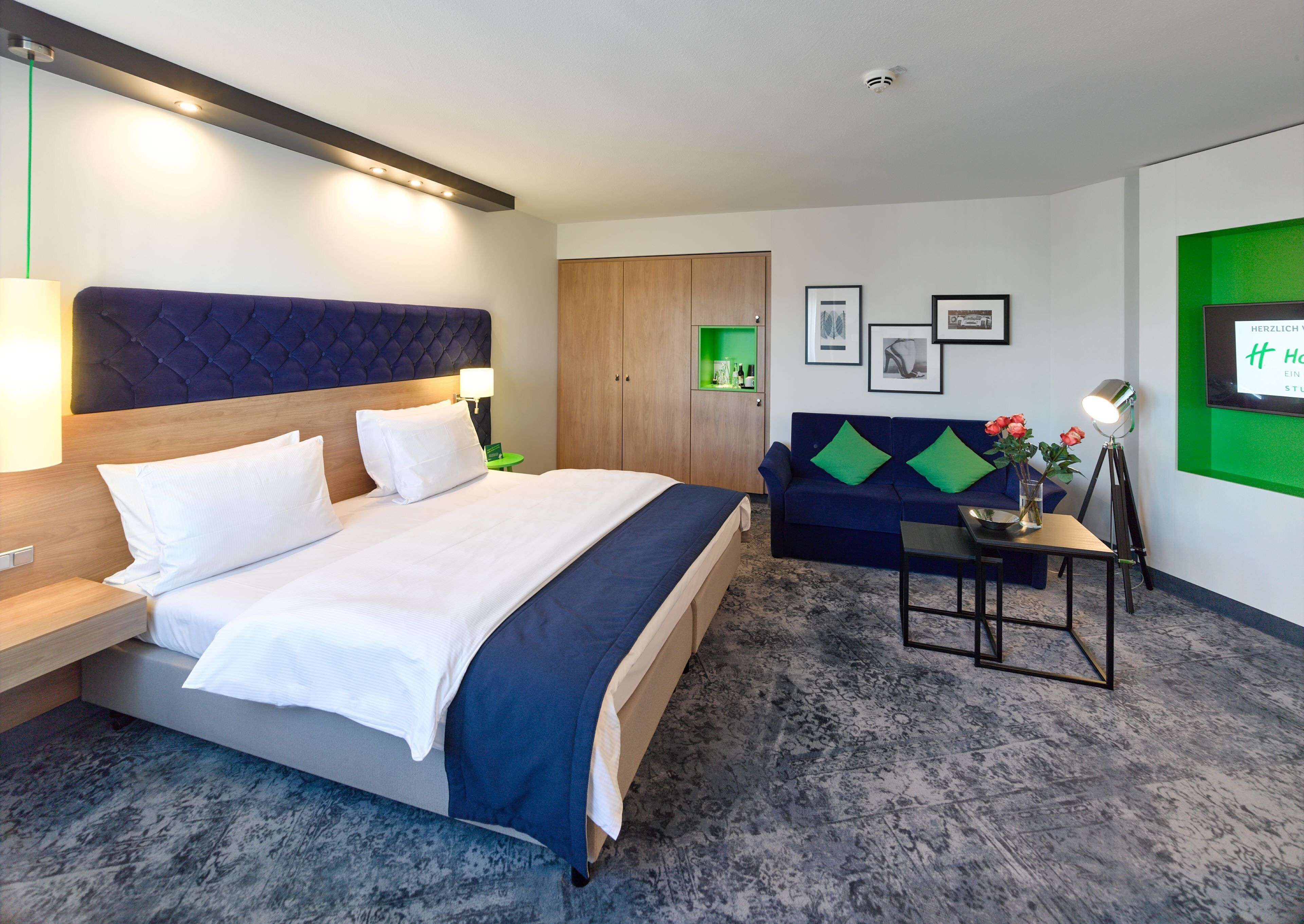 Une chambre d’hôtel avec un grand lit, un canapé, une petite table sur laquelle est posé un vase de fleurs, et un mur d’accent de couleur verte.