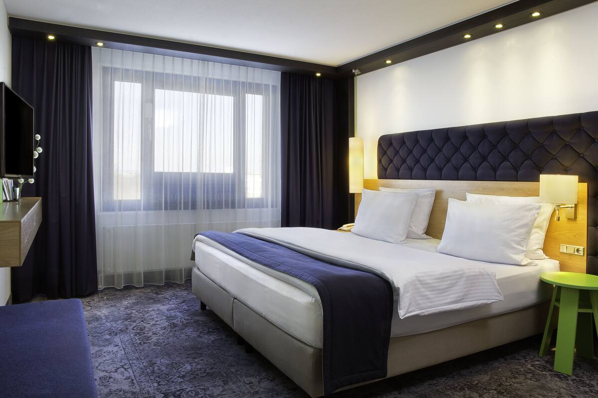 A hotel room with a large bed, a desk with a flat-screen TV, a green stool, and a window with sheer curtains.