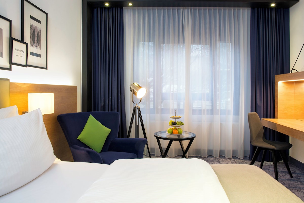 Hotellihuone, jossa on sänky, työpöytä, tuoli, lamppu ja pieni pöytä, jonka päällä on hedelmiä.