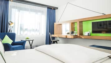 A hotel room with a bed, a desk with a chair, a desk lamp, a coffee maker, and a TV.