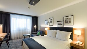 A hotel room with a large bed, a desk, a chair, a small table, and a sofa. There are framed pictures on the wall and a lamp on the bedside table. - Holiday Inn Stuttgart by IHG (Stuttgart)