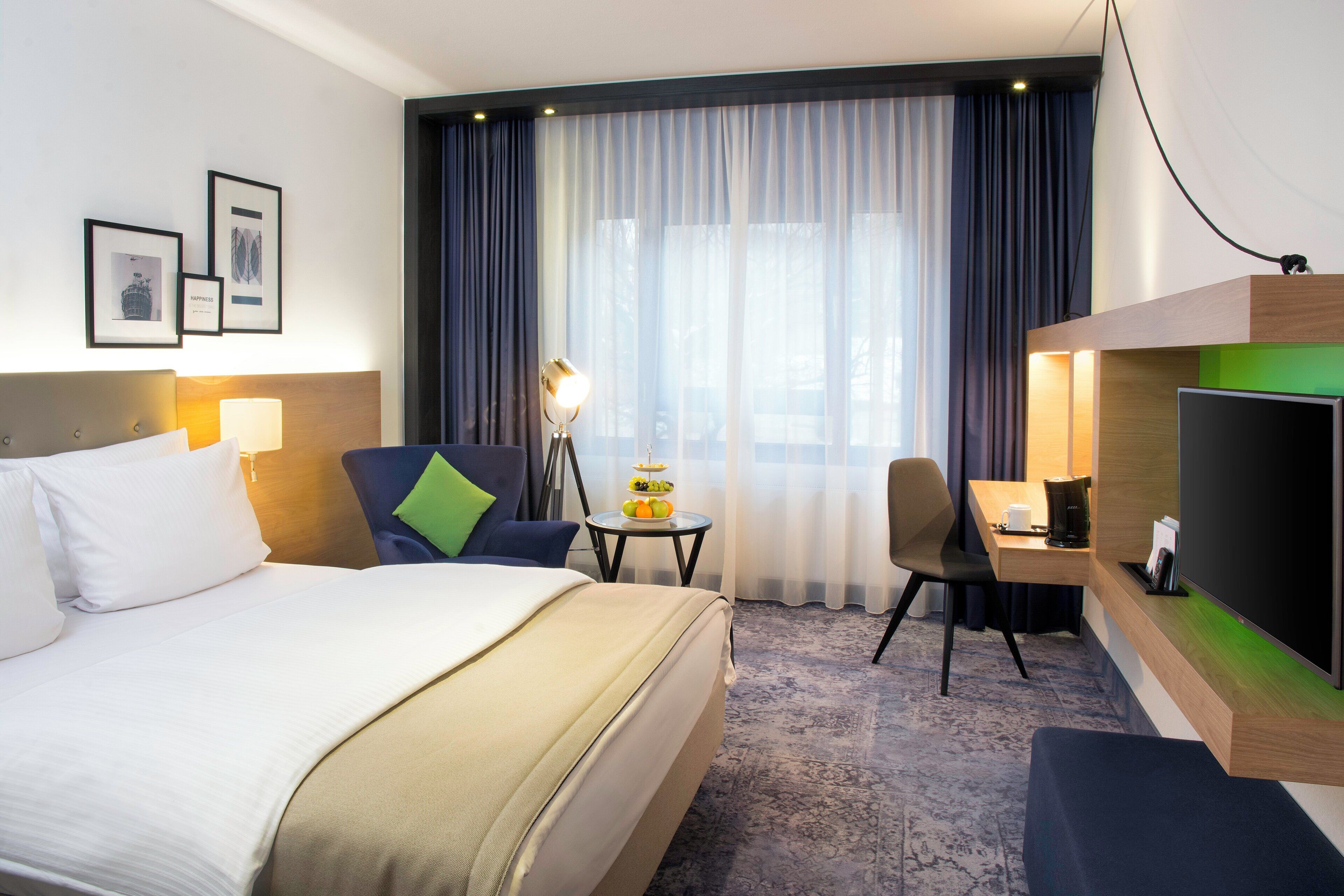 A hotel room with a bed, a desk, a chair, a lamp, a TV, and a window with curtains.