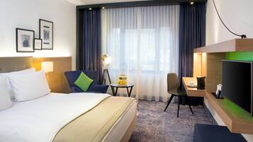 A hotel room with a bed, a desk, a chair, a lamp, a TV, and a window with curtains.