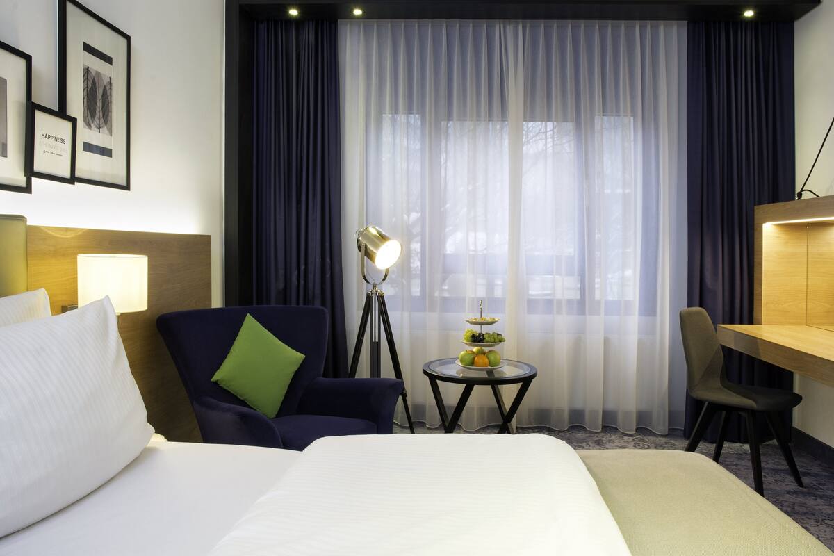 A hotel room with a bed, a desk, a chair, a lamp, and a small table with fruit.