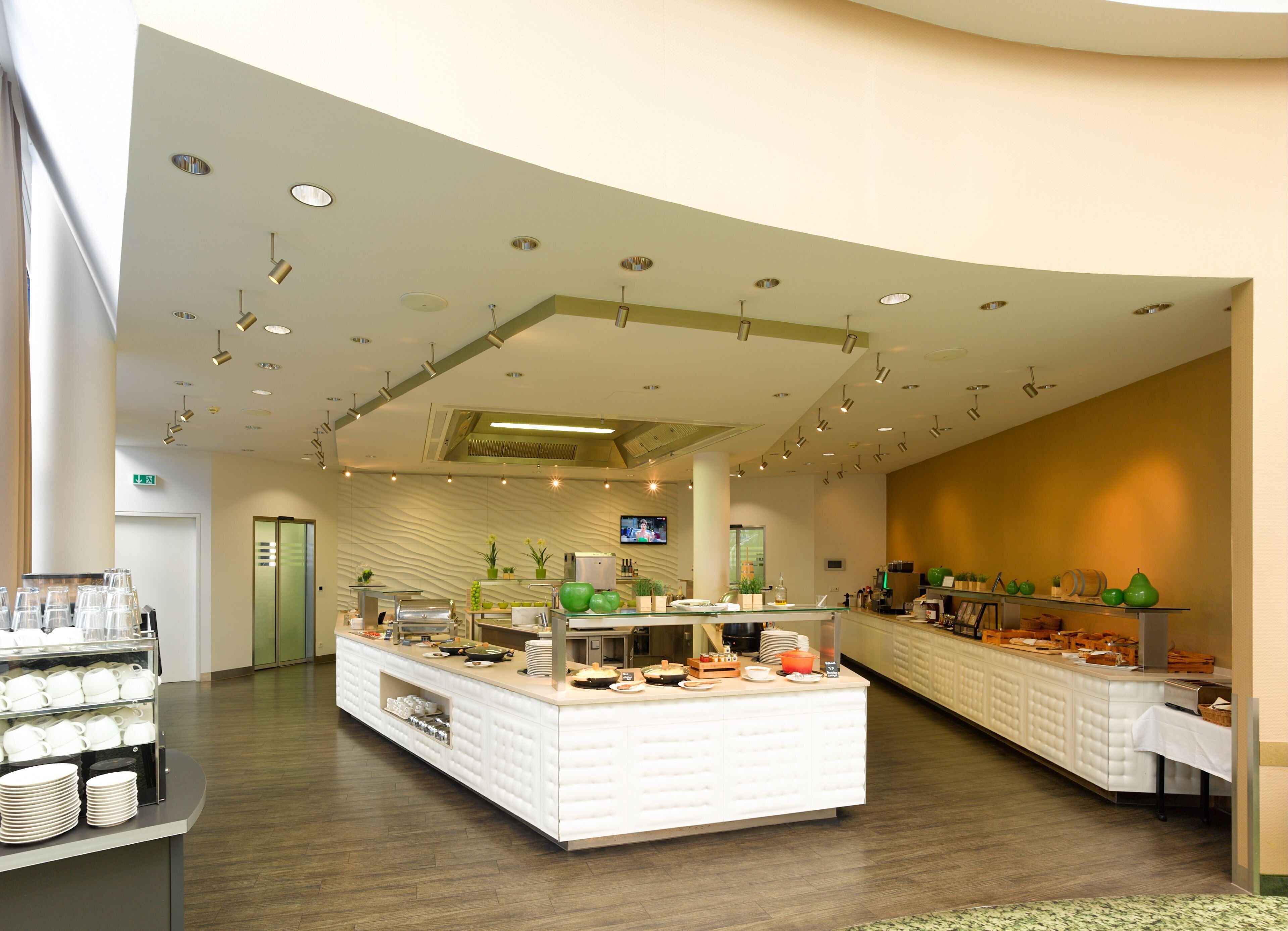 A modern hotel lobby with a central buffet area, a flat-screen TV mounted on the wall, and a green exit sign.