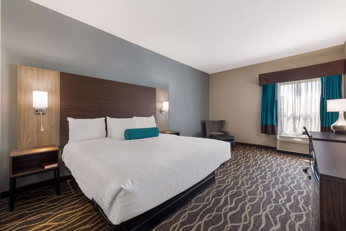 A hotel room with a large bed, a wooden headboard, a nightstand with a lamp, a desk with a lamp, and a window with curtains.