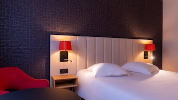 A hotel room with a bed, two red wall lamps, a red chair, and a small wooden side table.