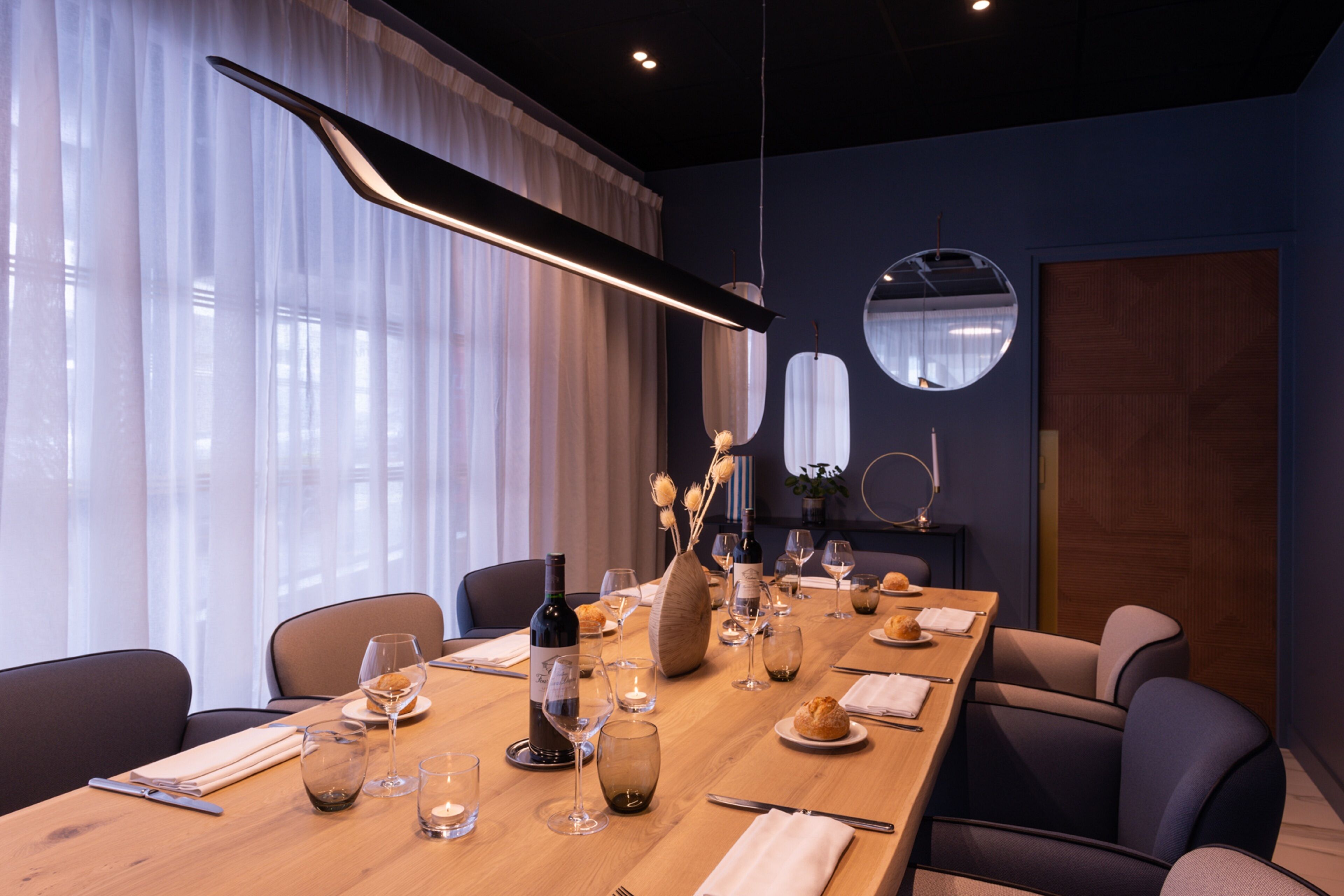 A modern dining room with a long table set for a meal, featuring wine glasses, plates, and a bottle.