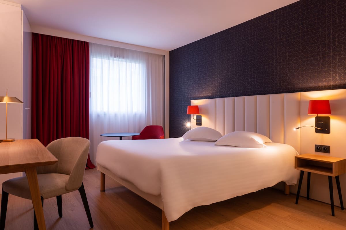 A hotel room with a bed, a desk, a chair, and a red curtain.