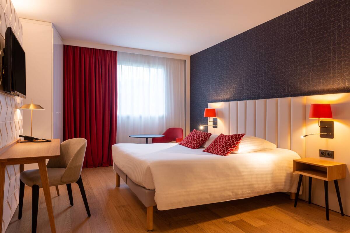 A hotel room with a bed, a desk, a chair, a TV, and a window with curtains.