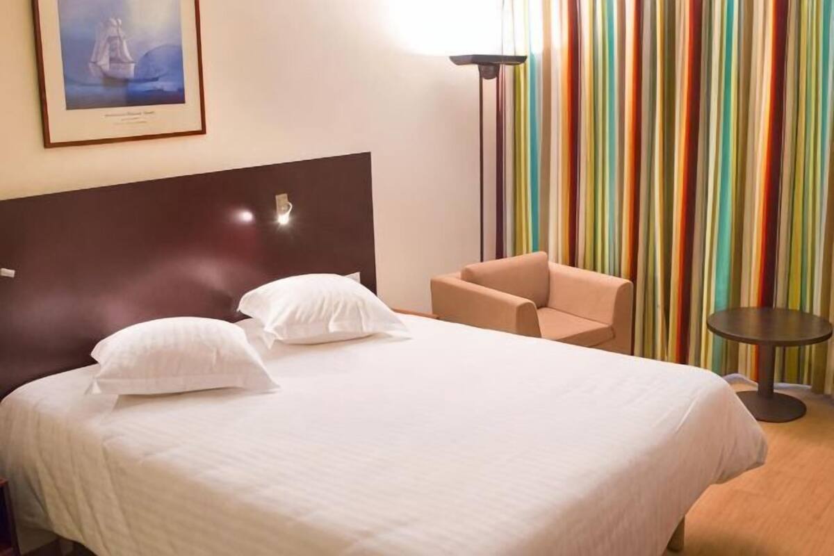 A hotel room with a bed, a chair, a small table, and a colorful striped curtain.