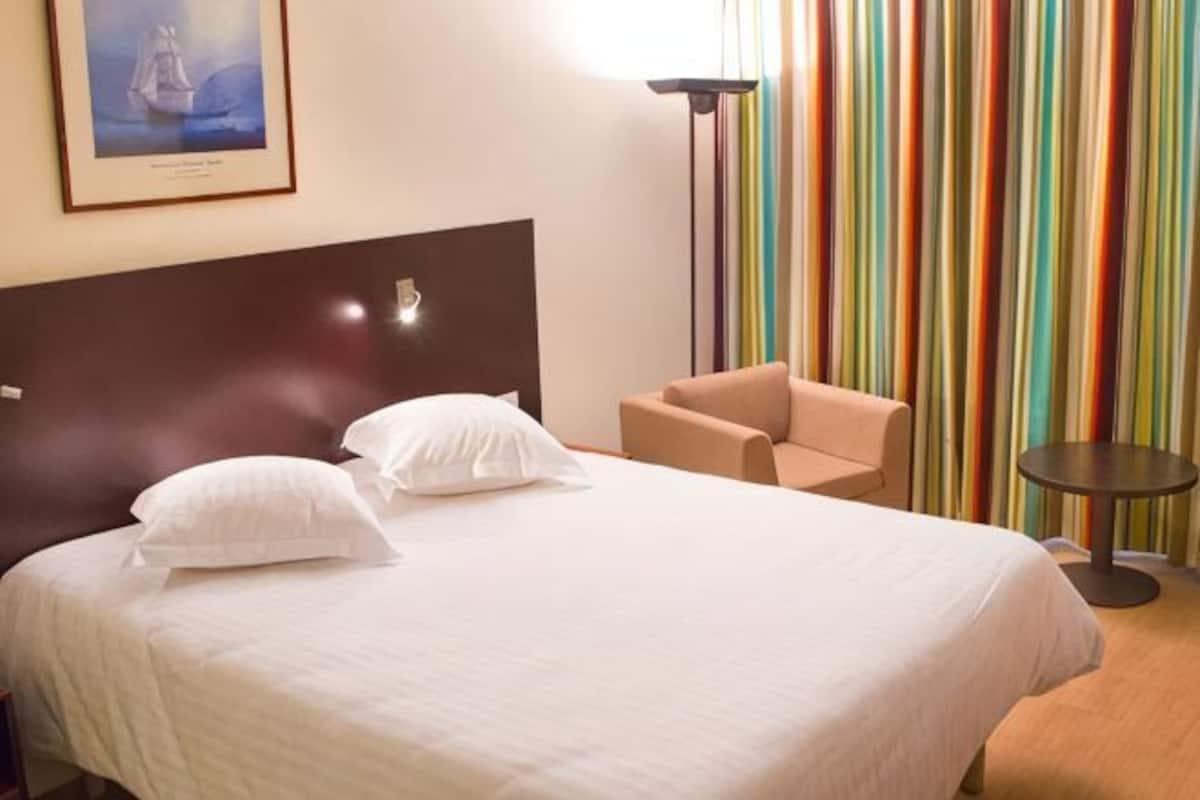 Quarto de hotel com cama, uma cadeira, uma mesinha e cortina listrada colorida.