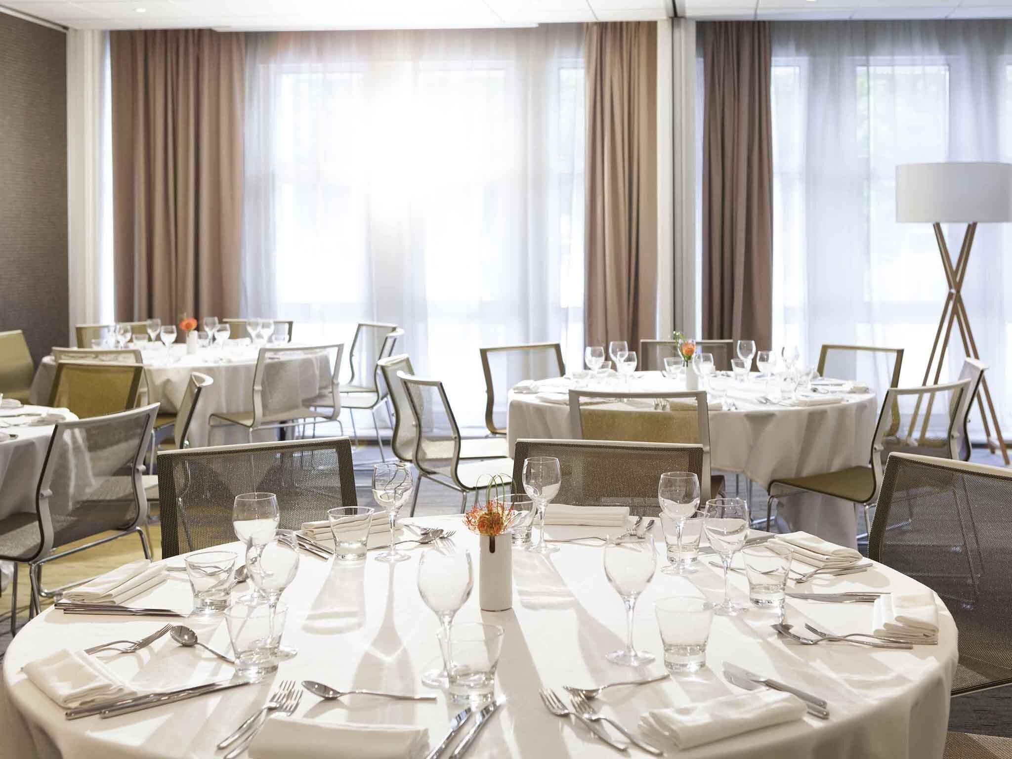 A hotel dining area with round tables set for a meal, large windows with curtains, and a modern chandelier.