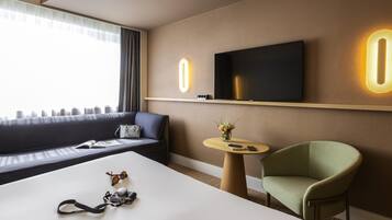 Una habitación de hotel moderna con una cama grande, un sofá, un televisor montado en la pared, una pequeña mesa con un florero y un sillón verde.