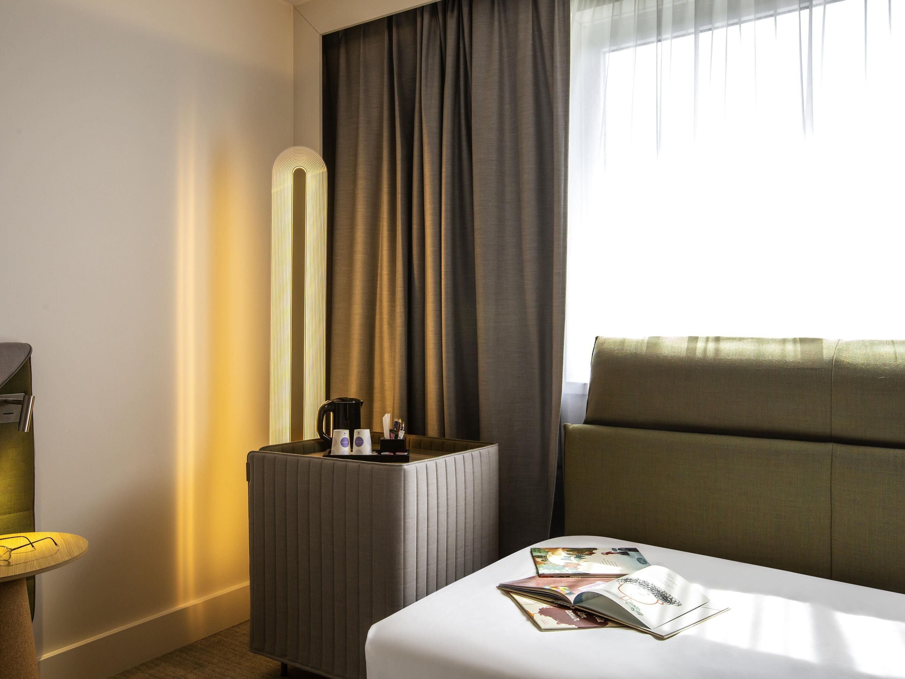 Una habitación de hotel con cama, una mesita de noche con lámpara, cafetera y una revista.
