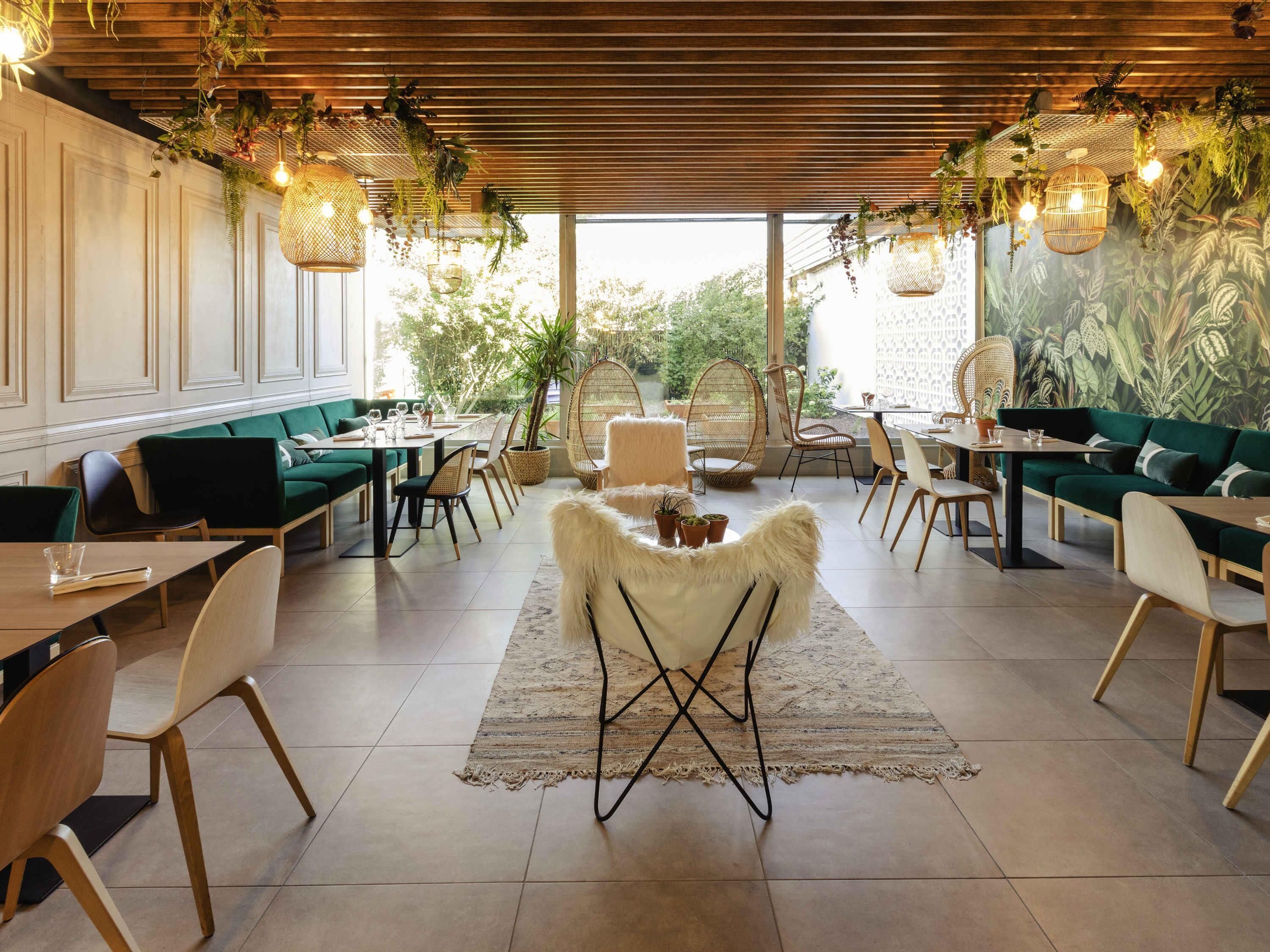 Un comedor moderno con sofás tapizados en verde, mesas de madera, sillas de mimbre y plantas colgantes.