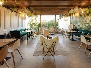 A modern dining area with green upholstered sofas, wooden tables, wicker chairs, and hanging plants. - Novotel Nancy (Laxou)