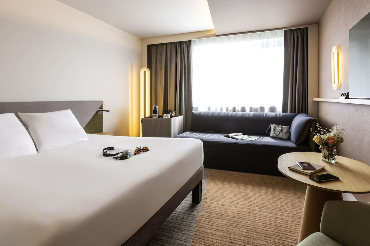 A hotel room with a bed, a sofa, a small table, a lamp, and a TV.