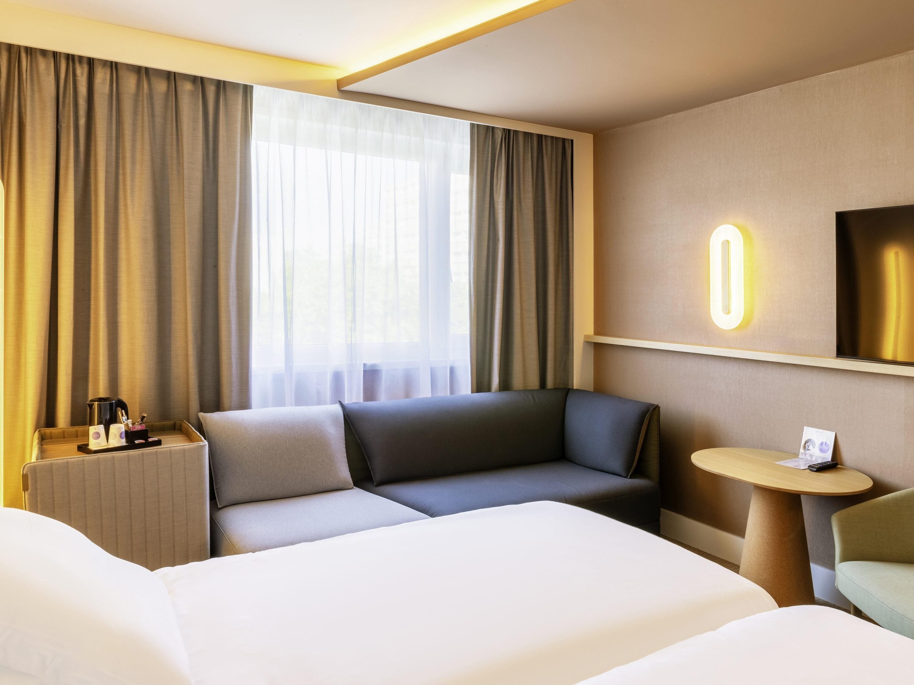 A hotel room with a bed, a sofa, a small table, a nightstand, and a TV.