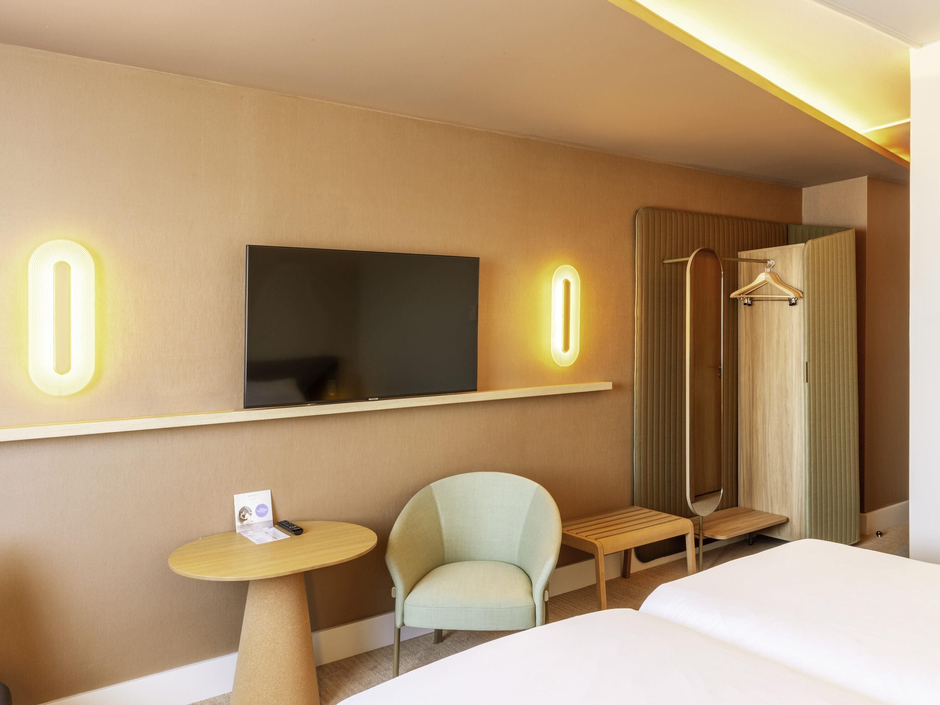 Camera d'albergo con un letto, una sedia, un tavolino, una TV e uno specchio con numeri illuminati.
